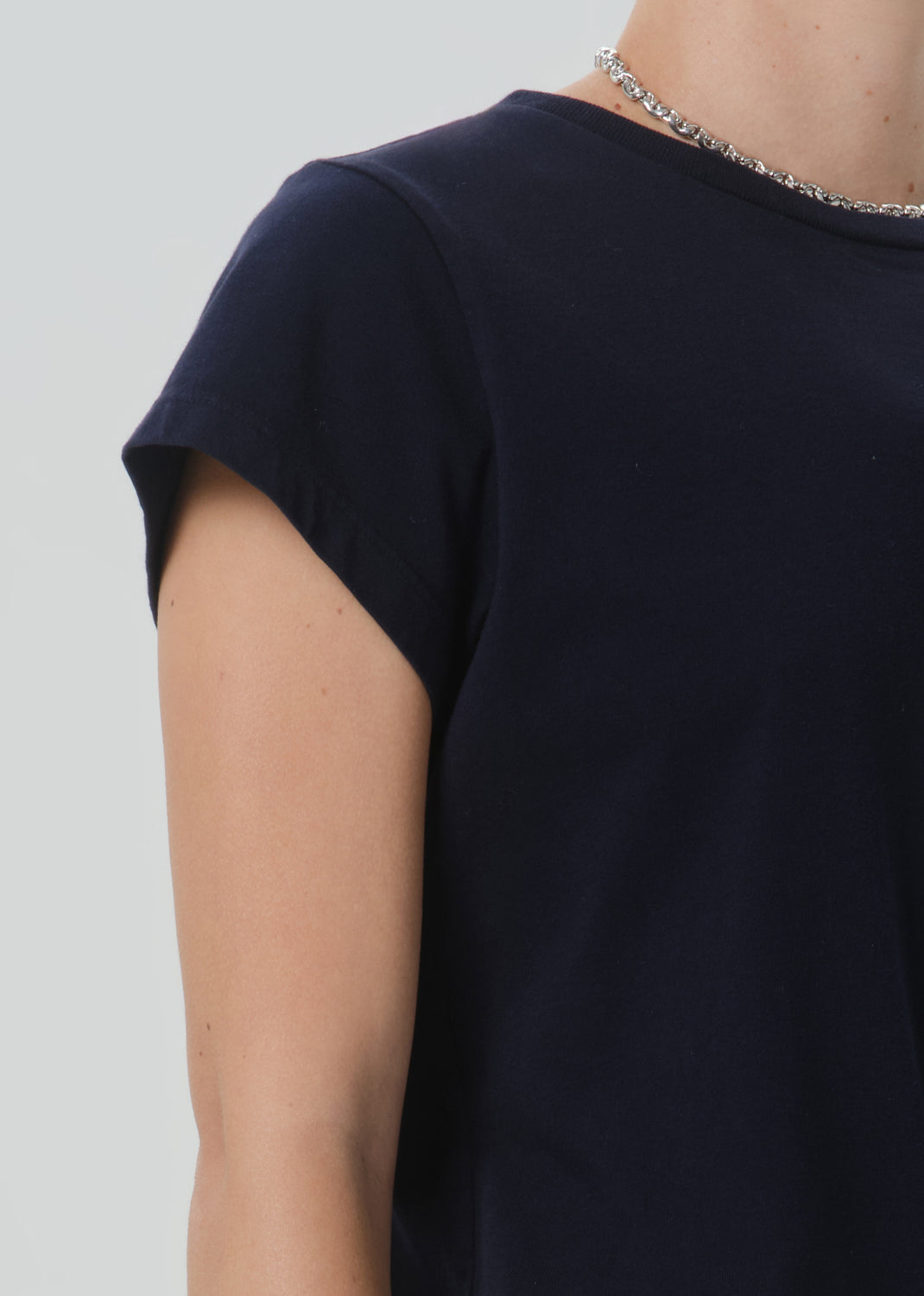 Close-up of a person wearing a dark navy short-sleeve t-shirt and a silver chain necklace against a grey background.