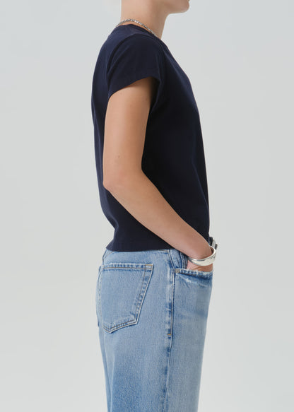 A side-profile view of a person wearing a dark navy short-sleeve crew-neck t-shirt and light-wash blue jeans against a grey background.