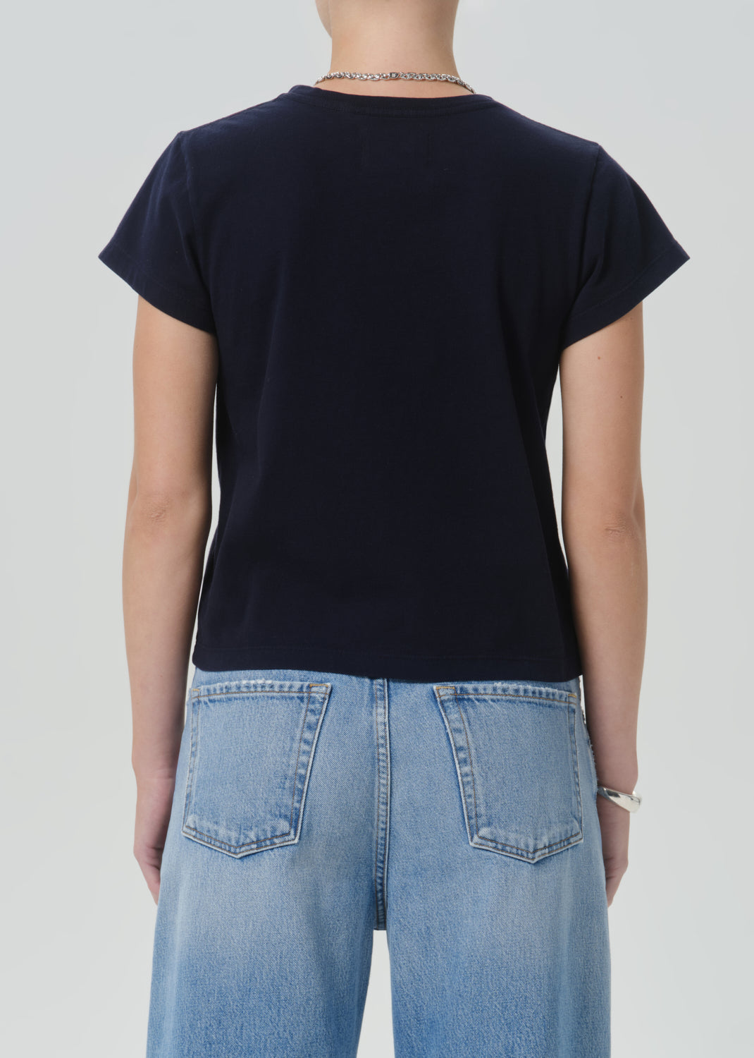 A person shown from the back wearing a dark navy short-sleeve crewneck T-shirt and light blue jeans against a grey background.