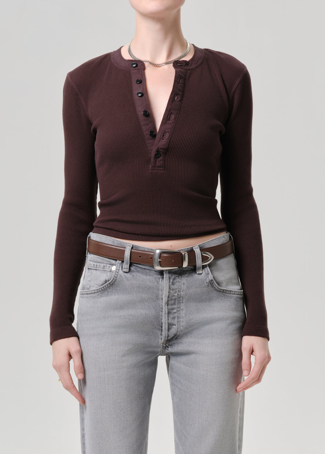 A model wearing a dark brown ribbed long-sleeve henley top with a button placket, paired with light grey jeans and a brown belt, against a grey background.