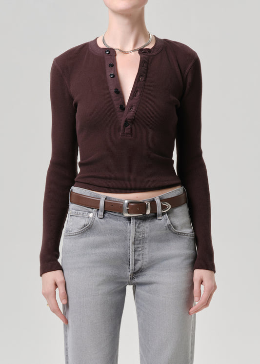 A model wearing a dark brown ribbed long-sleeve henley top with a button placket, paired with light grey jeans and a brown belt, against a grey background.