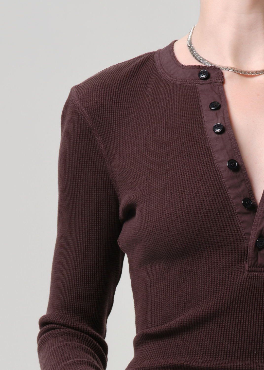 A close-up of a person wearing a dark brown waffle-knit long-sleeve henley top with black buttons, paired with a silver chain necklace, against a grey background.