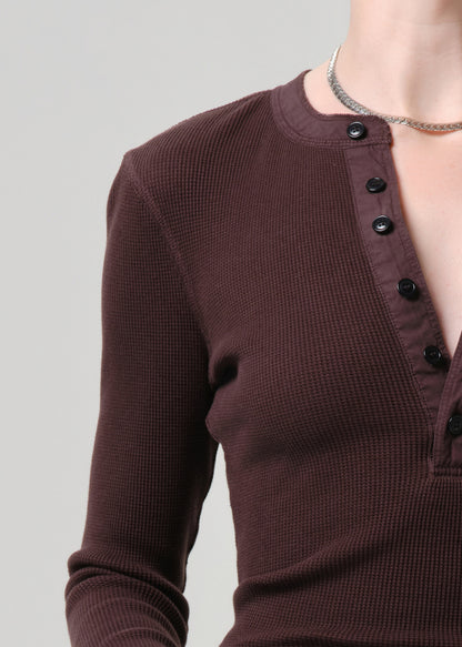 A close-up of a person wearing a dark brown waffle-knit long-sleeve henley top with black buttons, paired with a silver chain necklace, against a grey background.