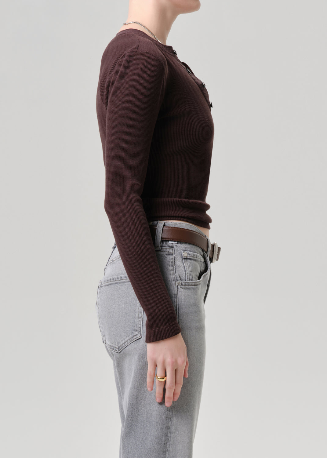 Side profile of a model wearing a dark brown long-sleeve ribbed knit top tucked into light grey jeans with a brown belt, against a grey background.