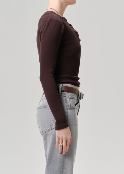 Side profile of a model wearing a dark brown long-sleeve ribbed knit top tucked into light grey jeans with a brown belt, against a grey background.