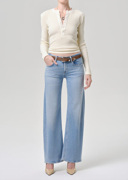 A model wearing a cream long-sleeve henley top with lace trim, light-wash wide-leg jeans, a brown belt, and beige heels, photographed against a grey background.