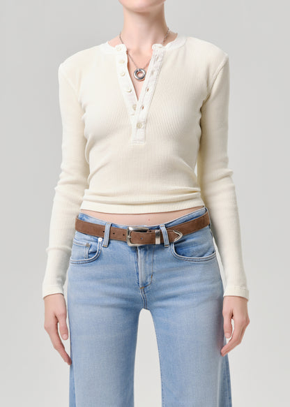 A model wearing a cream ribbed long-sleeve henley top tucked into light-wash blue jeans with a brown belt, shown from the neck down against a grey background.