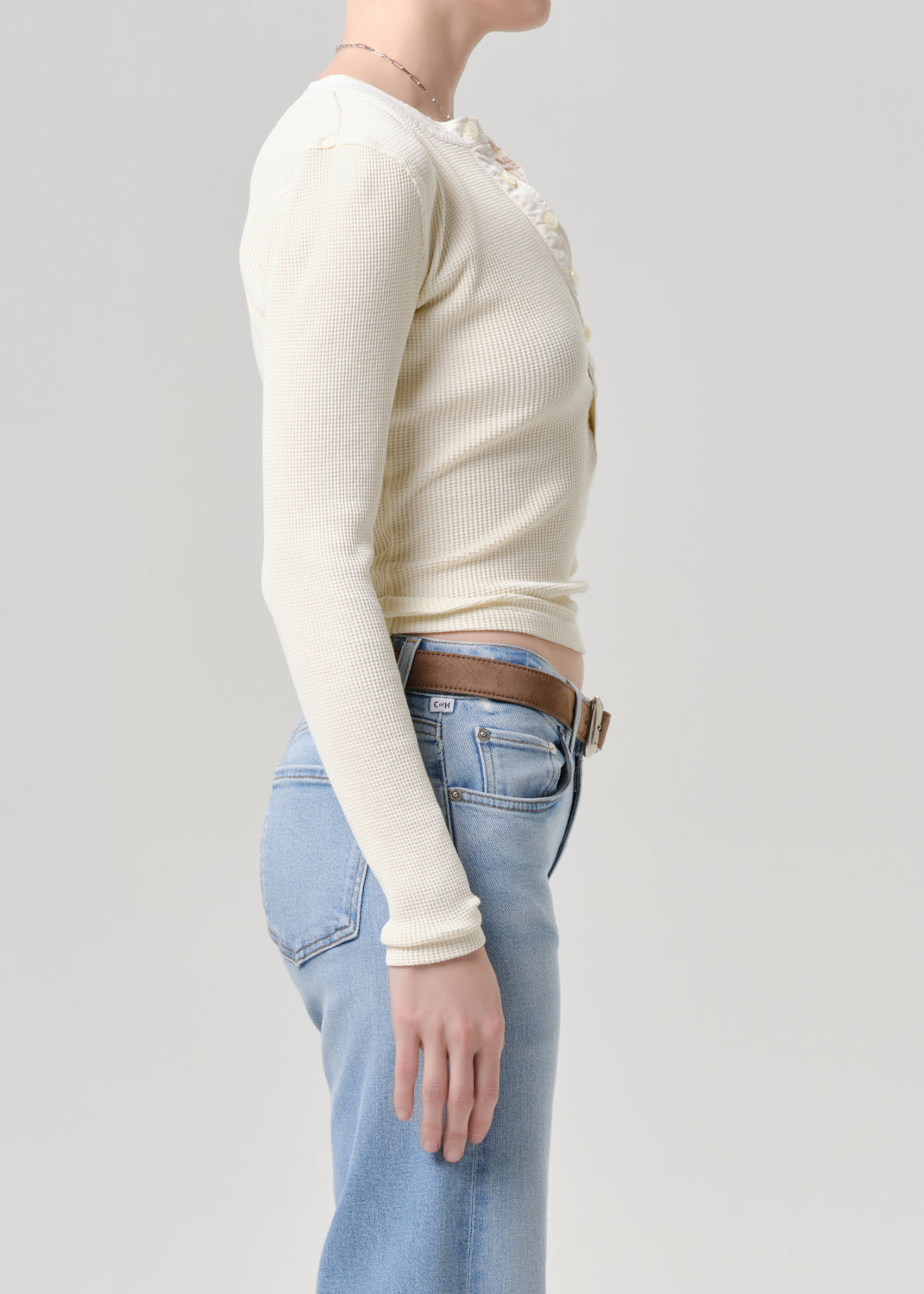 A side-profile view of a model wearing a cream ribbed long-sleeve cropped top with decorative buttons, paired with light blue jeans and a brown belt, against a grey background.