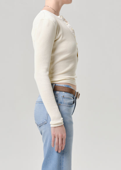 A side-profile view of a model wearing a cream ribbed long-sleeve cropped top with decorative buttons, paired with light blue jeans and a brown belt, against a grey background.