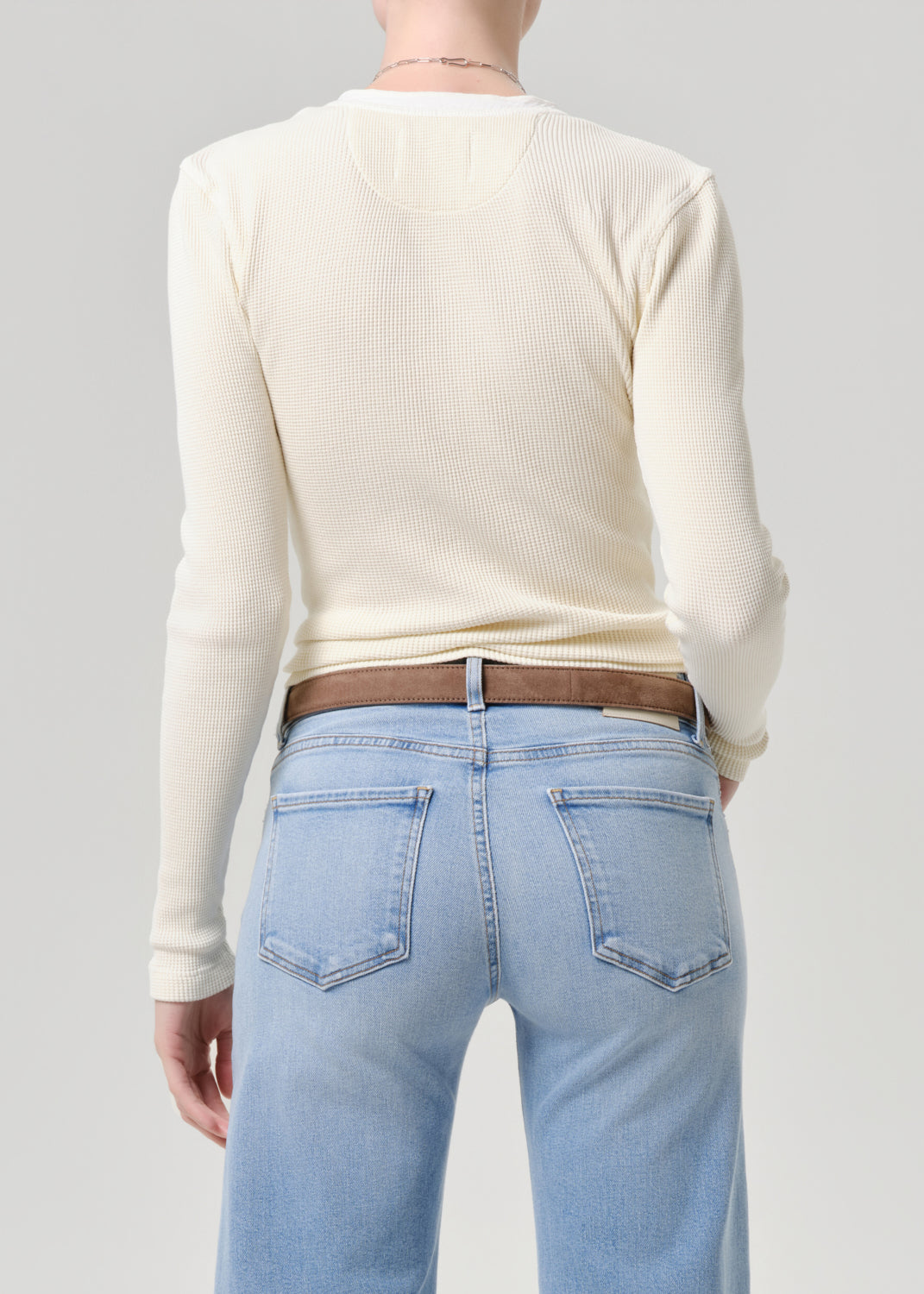 Back view of a person wearing a cream ribbed long-sleeve top, light blue jeans, and a brown belt against a grey background.