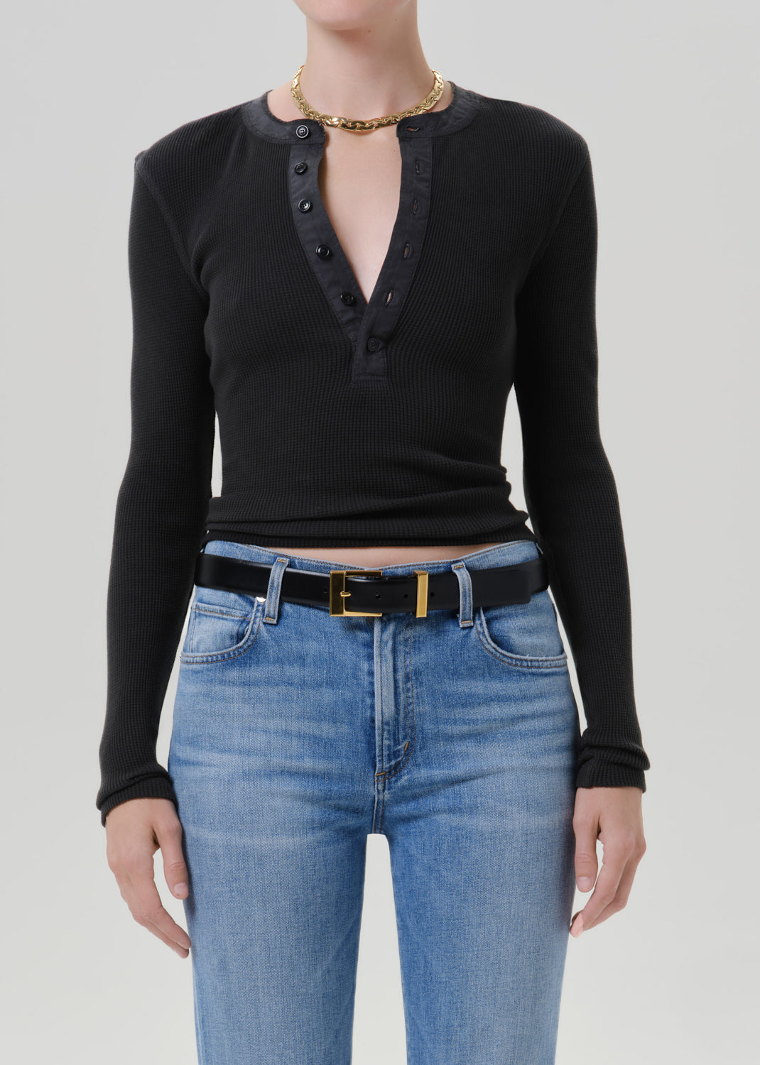 Person wearing a black long sleeve fitted henley top with light blue jeans and a black belt with gold buckle, front cropped view on a grey background.