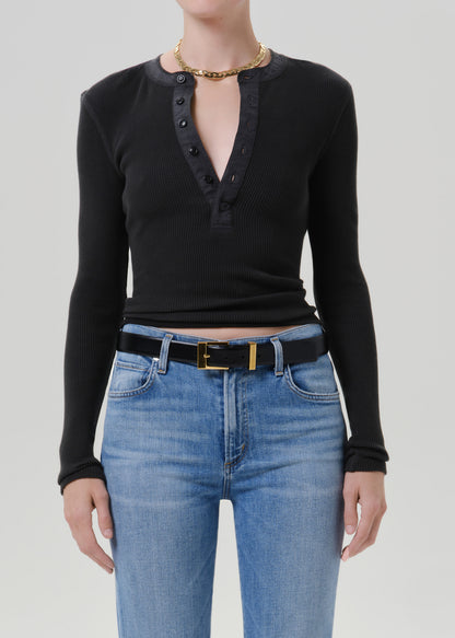 Person wearing a black long sleeve fitted henley top with light blue jeans and a black belt with gold buckle, front cropped view on a grey background.