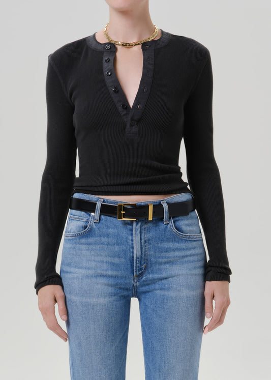 Person wearing a black long sleeve fitted henley top with light blue jeans and a black belt with gold buckle, front cropped view on a grey background.