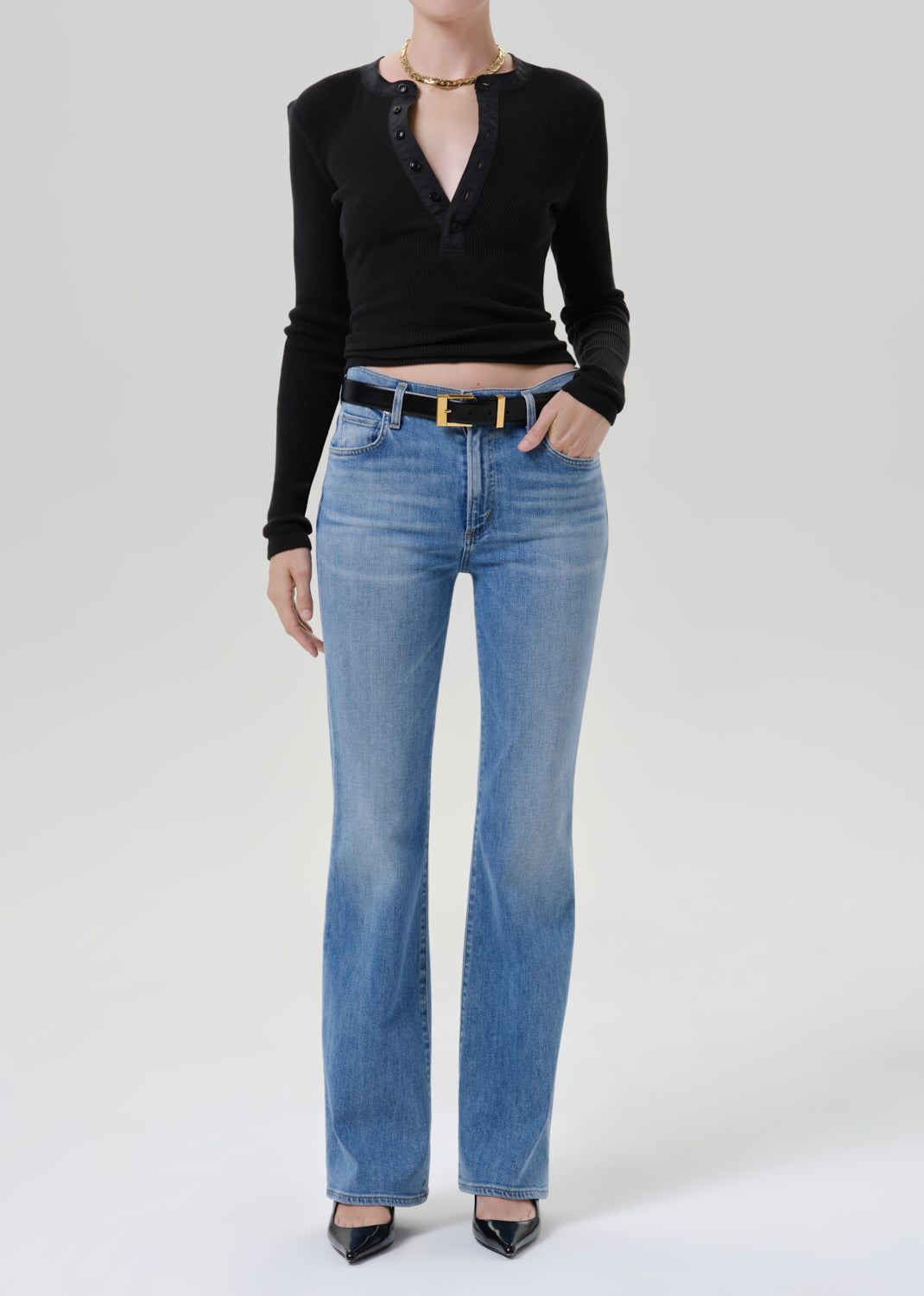 Person wearing a black long sleeve henley top with light wash straight leg jeans and black pointed toe heels on a grey background