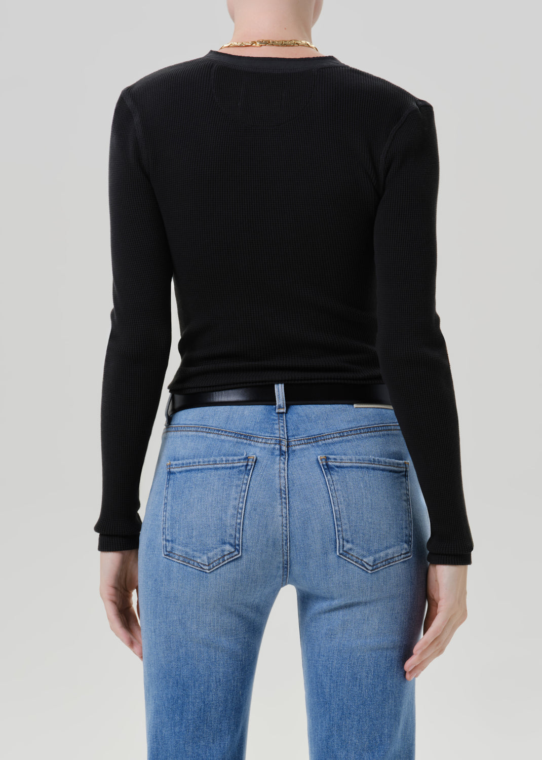 Person wearing a black long sleeve henley top with light wash straight leg jeans viewed from the back on a grey background