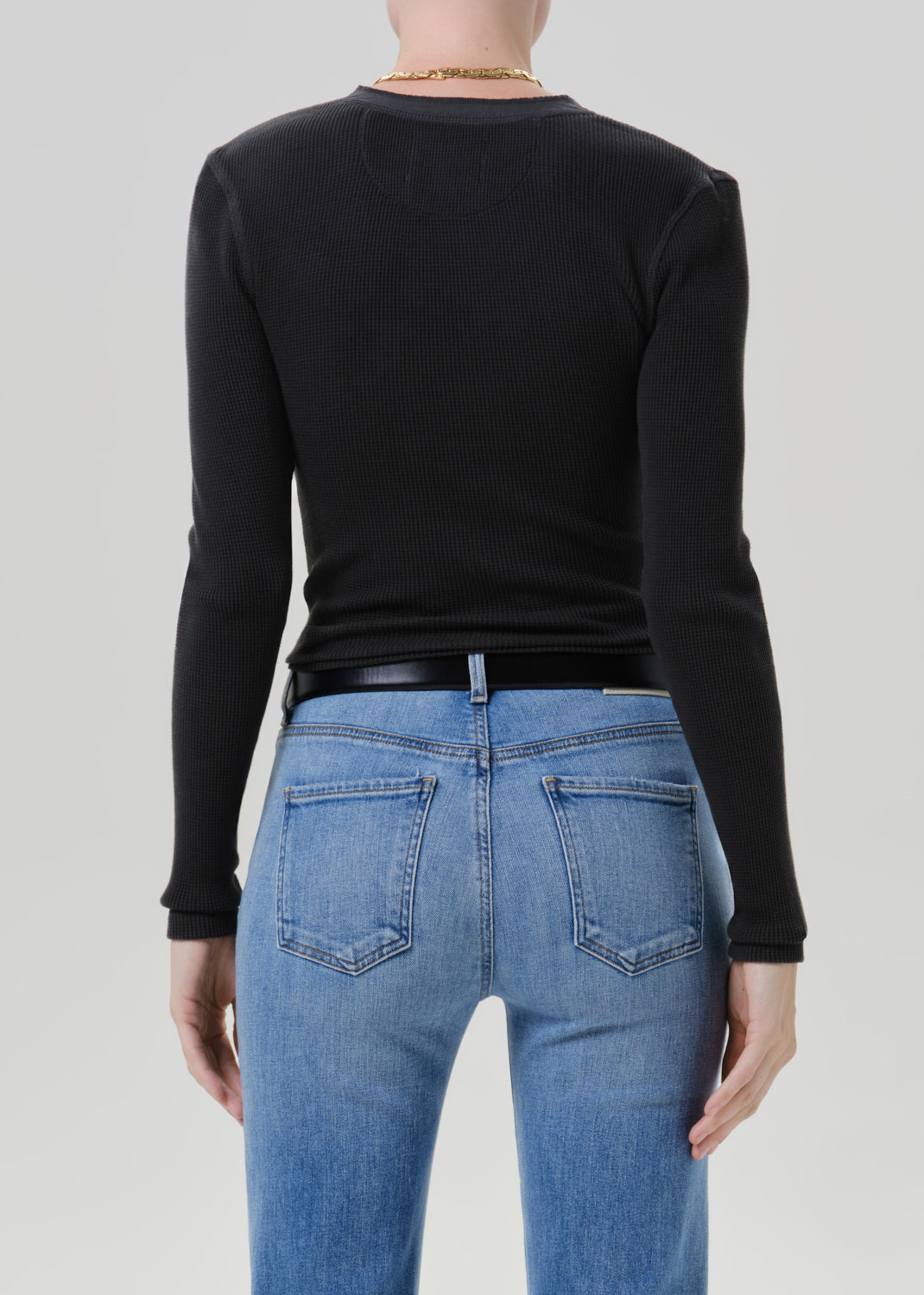 Person wearing a black long sleeve fitted henley top with light blue jeans, back cropped view on a grey background.