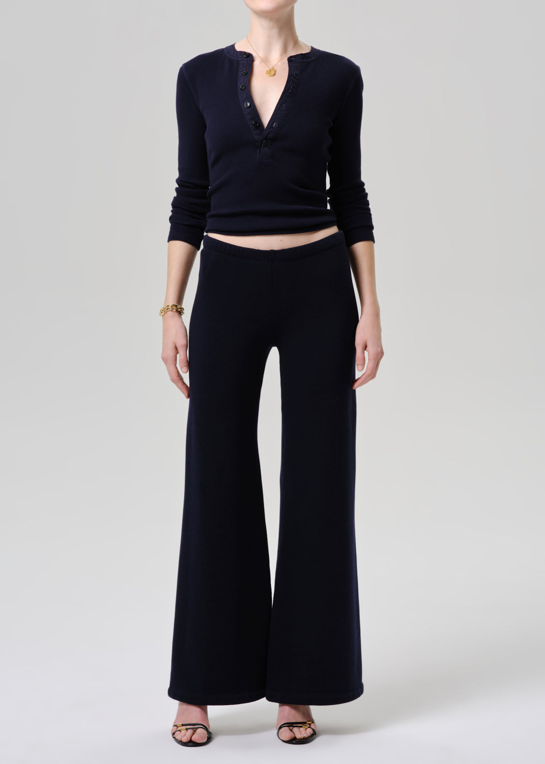 A model wearing a dark navy blue ribbed knit two-piece set consisting of a long-sleeve henley top with a deep V-neck and matching wide-leg trousers, styled with black sandals and a gold necklace on a grey background.