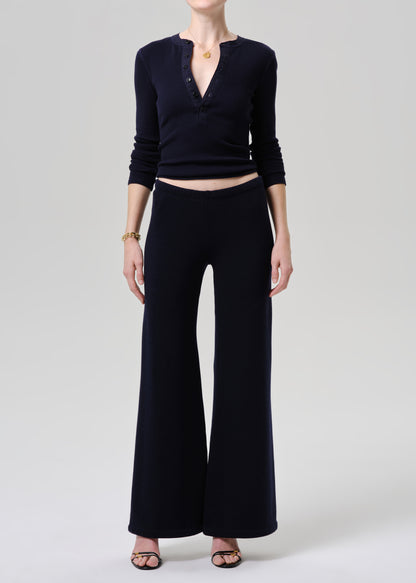 A model wearing a dark navy blue ribbed knit two-piece set consisting of a long-sleeve henley top with a deep V-neck and matching wide-leg trousers, styled with black sandals and a gold necklace on a grey background.