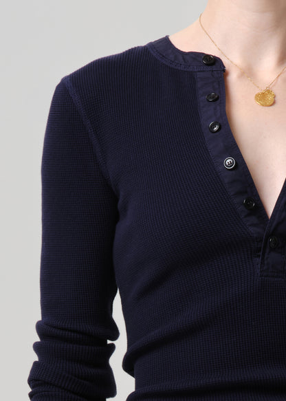 Close-up of a person wearing a dark navy ribbed long-sleeve henley top with black buttons, against a grey background.