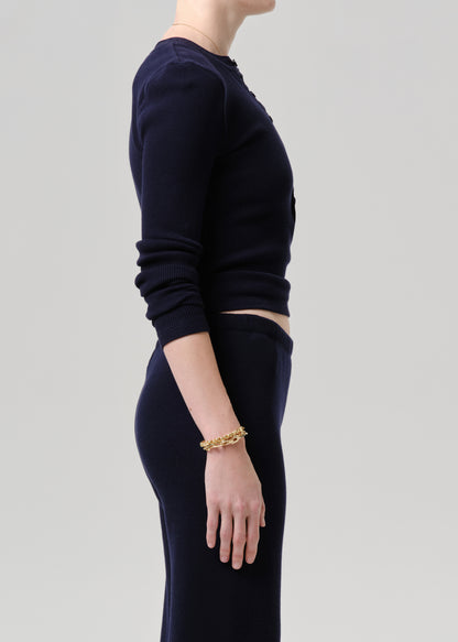 A model shown in side profile wearing a dark navy blue ribbed knit long-sleeve crop top and matching dark navy pants, accessorized with a chunky gold chain bracelet, against a grey background.