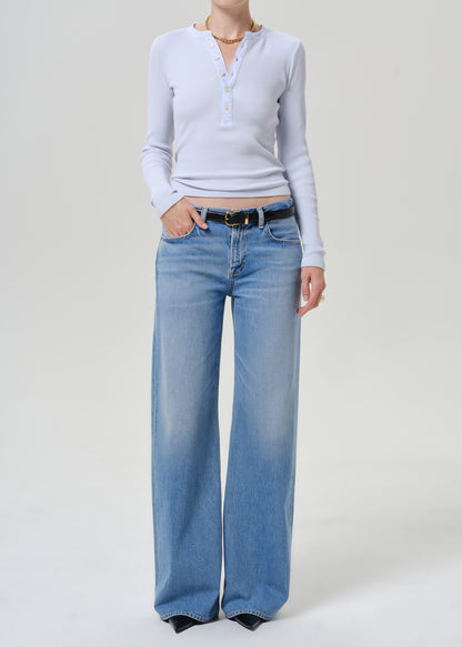 A model wearing a light blue long-sleeve henley top and wide-leg light blue jeans with a black belt, standing against a plain light gray studio background.