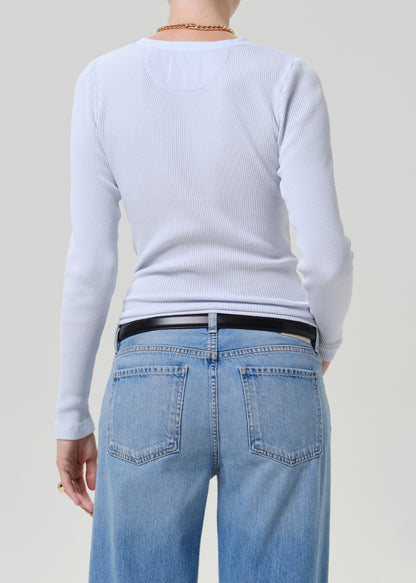 Back view of a person wearing a light blue ribbed long-sleeve top and light blue jeans with a black belt, against a plain light gray studio background.