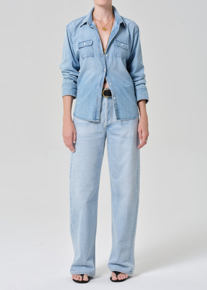 Person wearing a light blue denim shirt and jeans on a grey background