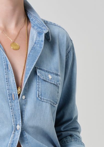 Close-up of a person wearing a blue denim shirt with a gold necklace on a plain background