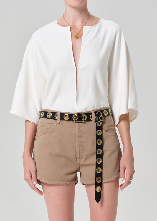 Person wearing a white blouse and beige shorts with a decorative belt on a plain background