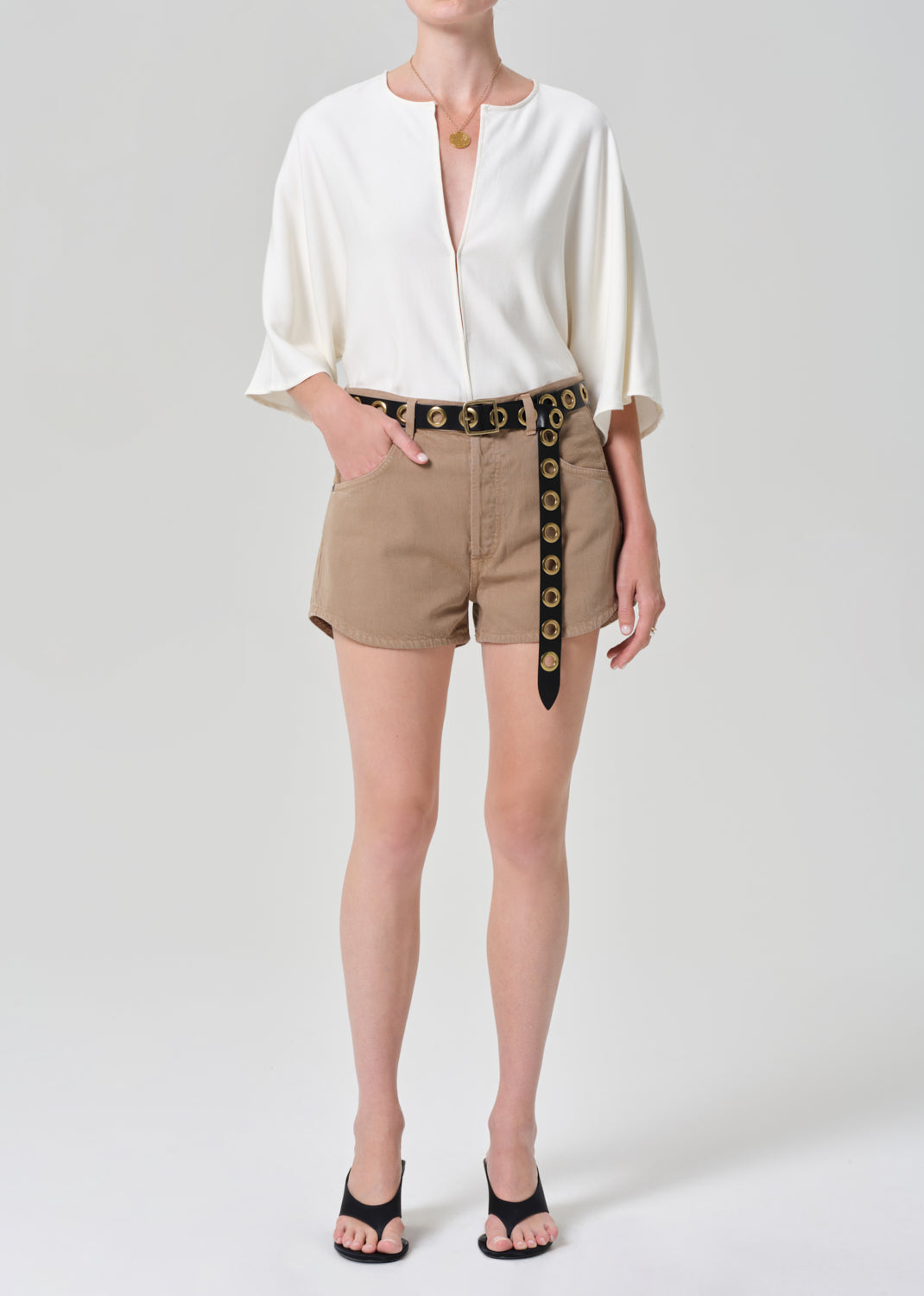 Person wearing a white blouse and beige shorts with a decorative belt on a plain background