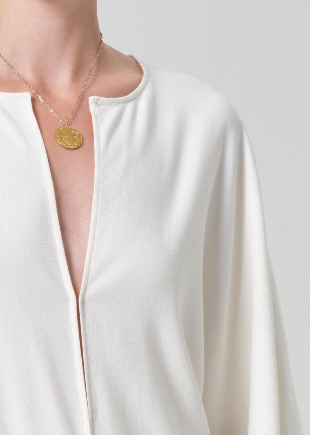Person wearing a white blouse and a gold necklace with a pendant on a plain background