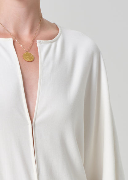 Person wearing a white blouse and a gold necklace with a pendant on a plain background