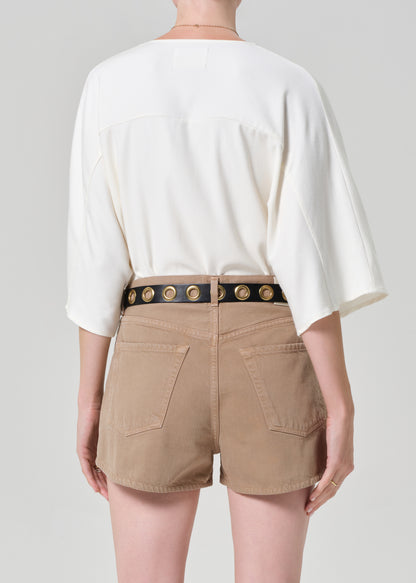 Person wearing a white blouse and beige shorts with a decorative belt on a plain background