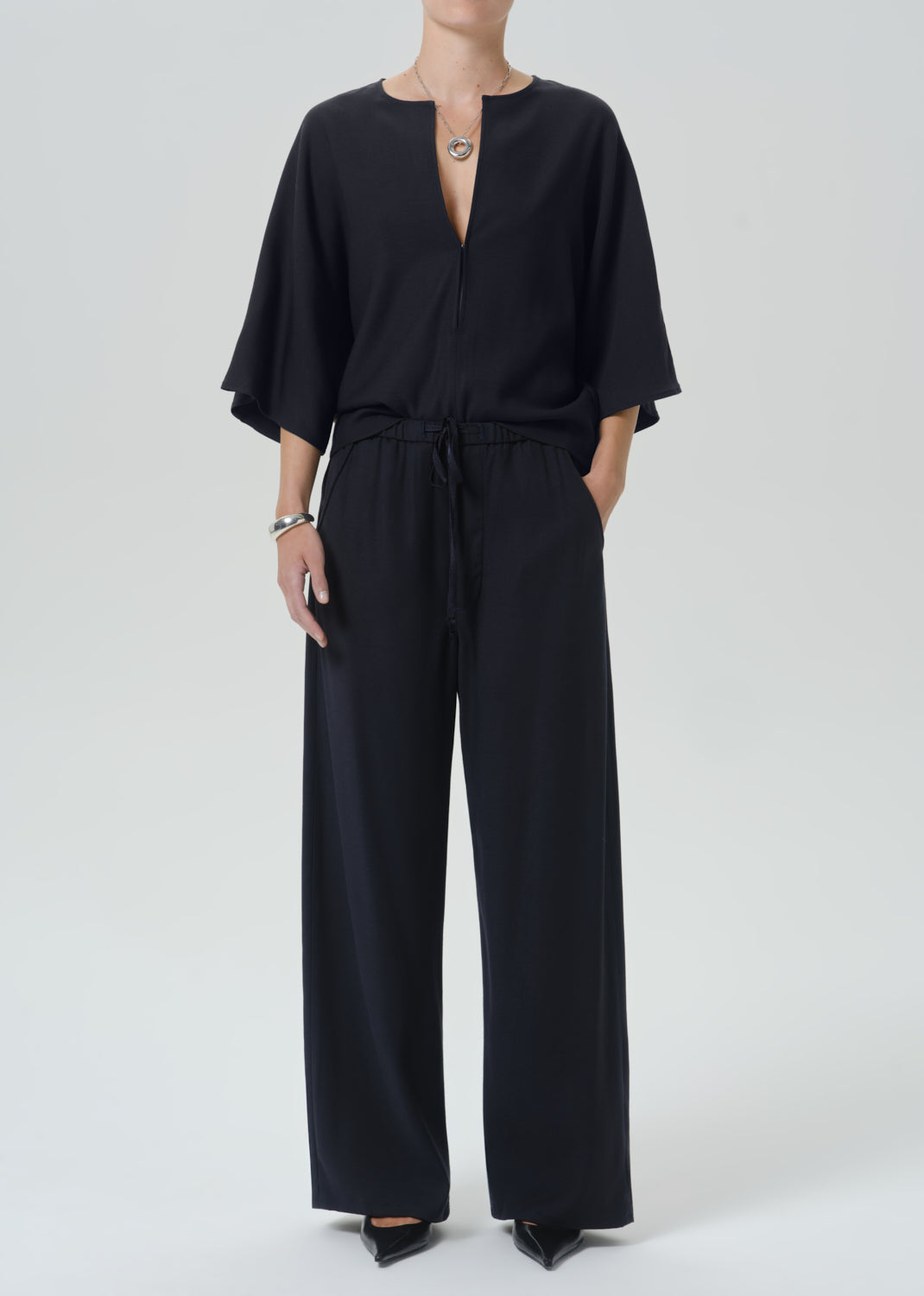 A model wearing a black wide-leg jumpsuit with a split neckline, short flutter sleeves, and a drawstring waist, photographed against a grey background.