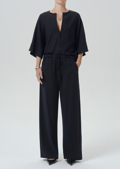 A model wearing a black wide-leg jumpsuit with a split neckline, short flutter sleeves, and a drawstring waist, photographed against a grey background.