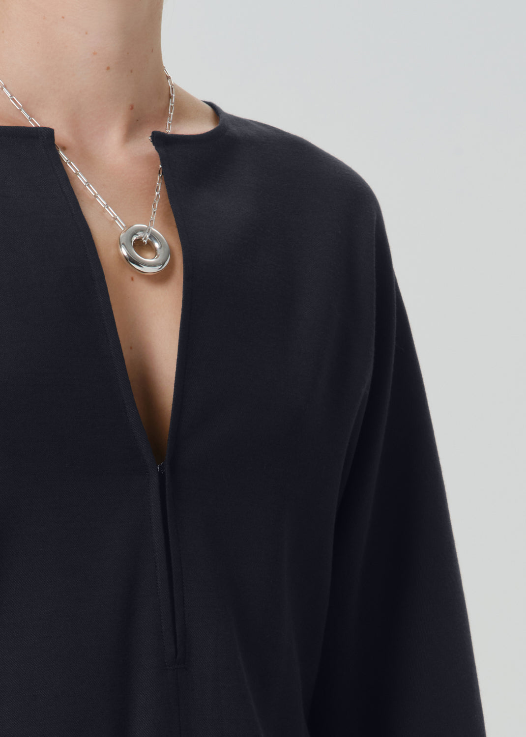 Close-up of a person wearing a silver-toned double-ring pendant necklace over a black top, against a grey background.