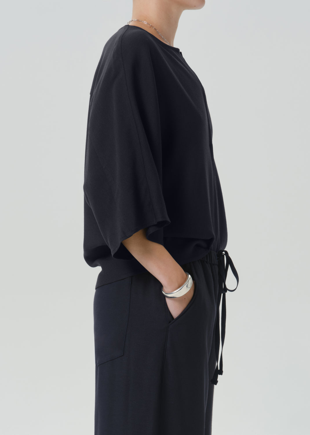 A side-profile view of a model wearing a black loose-fitting top with wide draped sleeves and matching black wide-leg trousers with a drawstring waist, set against a grey background.