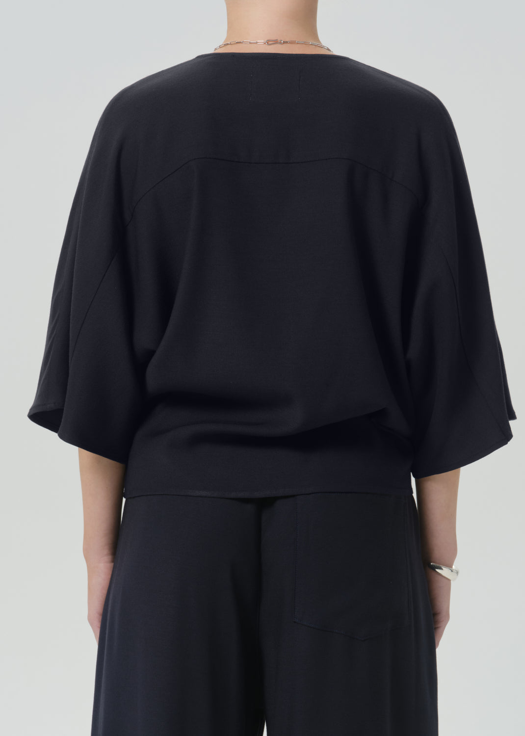 Rear view of a person wearing a loose black draped top with wide three-quarter sleeves and black wide-leg pants against a grey background.