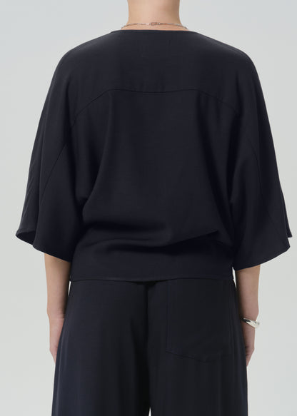 Rear view of a person wearing a loose black draped top with wide three-quarter sleeves and black wide-leg pants against a grey background.