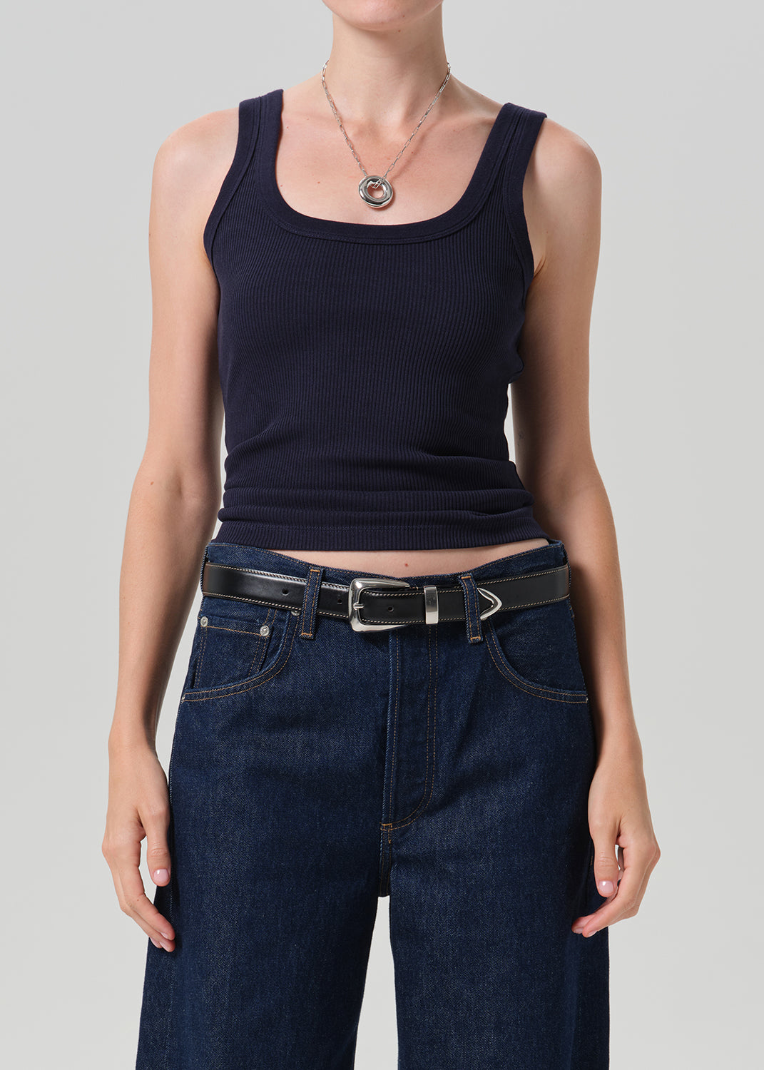 A model wearing a dark navy ribbed tank top tucked into dark blue jeans with a black belt, shot against a grey background.