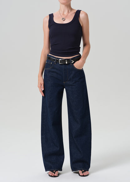 A model wearing dark blue wide-leg denim jeans with a black belt, paired with a black tank top and black sandals, photographed against a grey background.