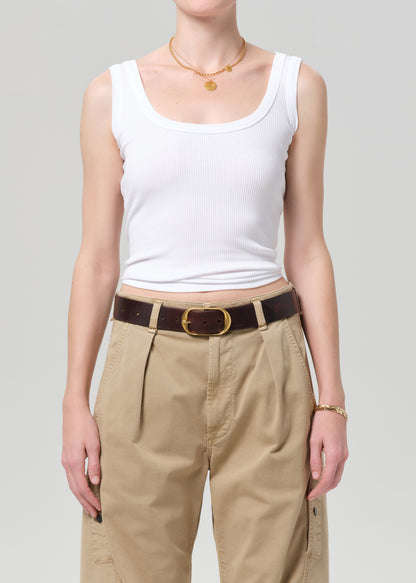 Person wearing a white tank top and beige pants with a brown belt on a plain background