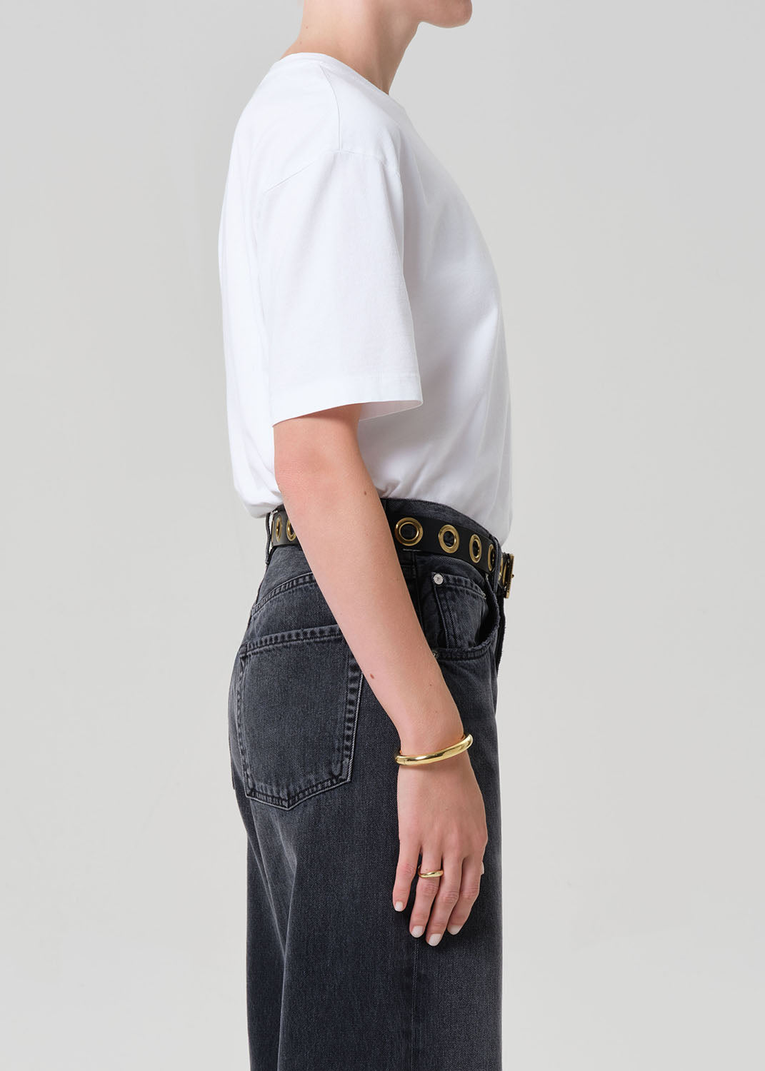 Person wearing a white t-shirt and dark jeans with a gold belt and bracelet on a light gray background