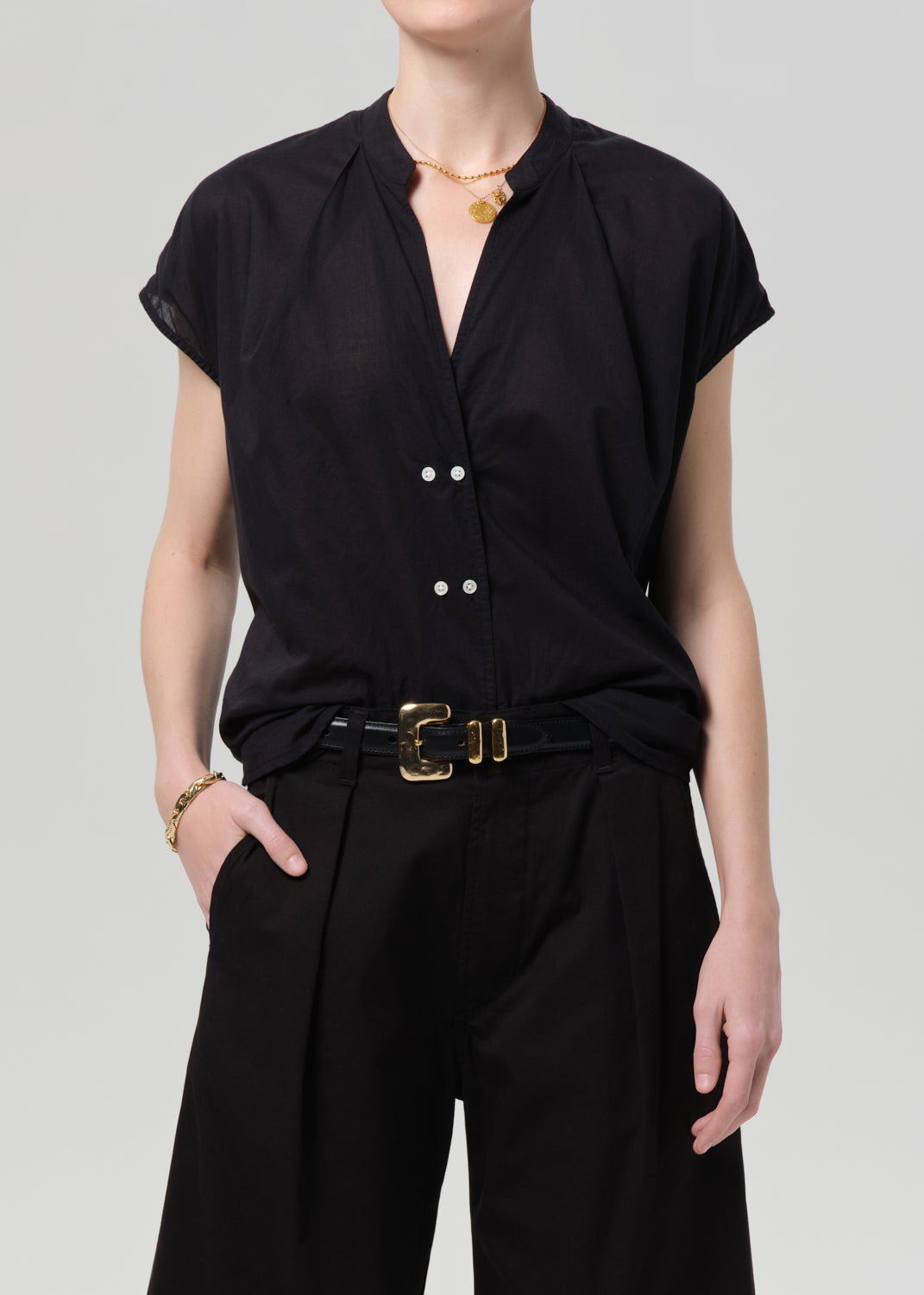 Person wearing a black blouse and black pants with a gold belt on a gray background