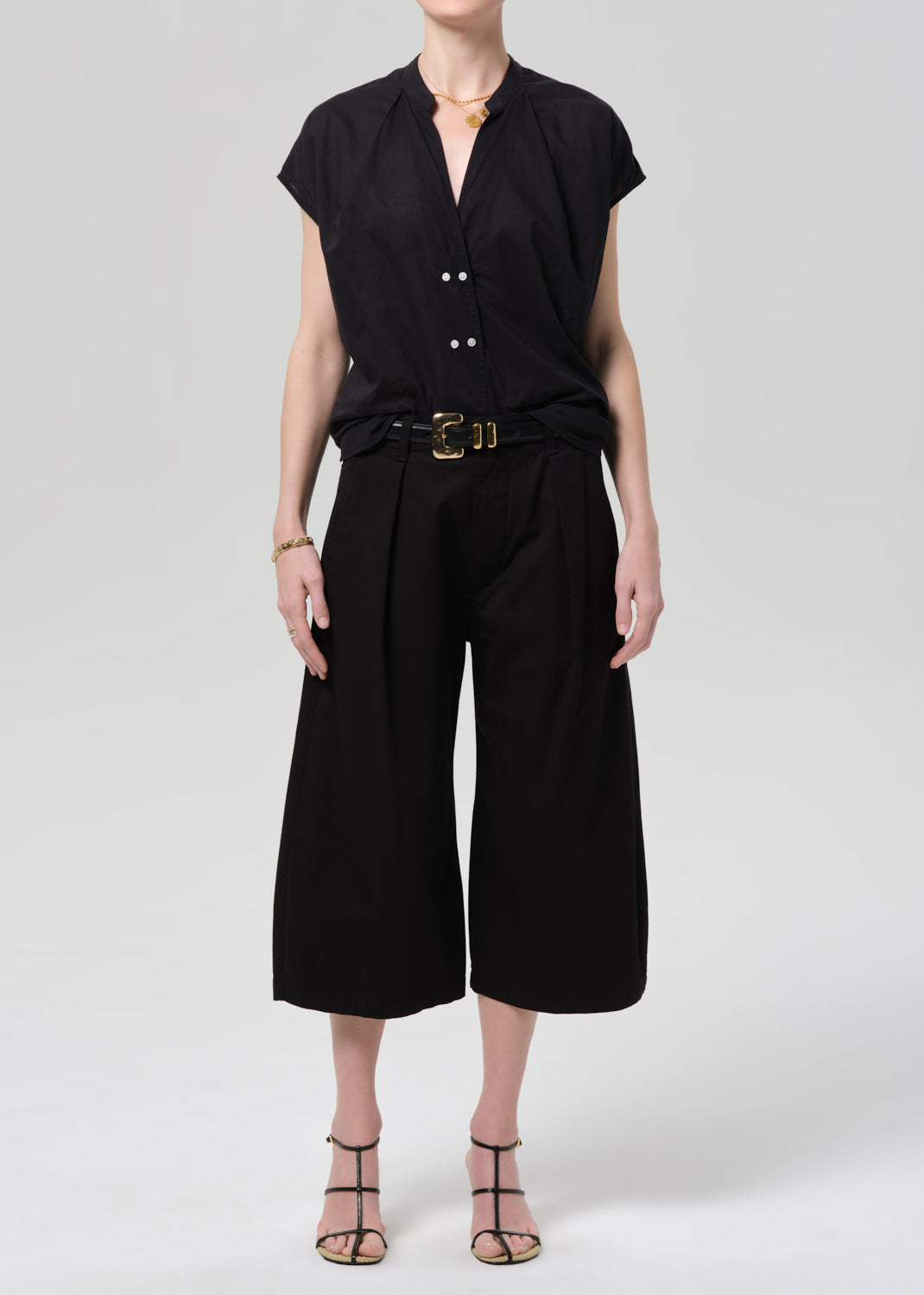 Person wearing a black blouse and black pants with a gold belt on a gray background