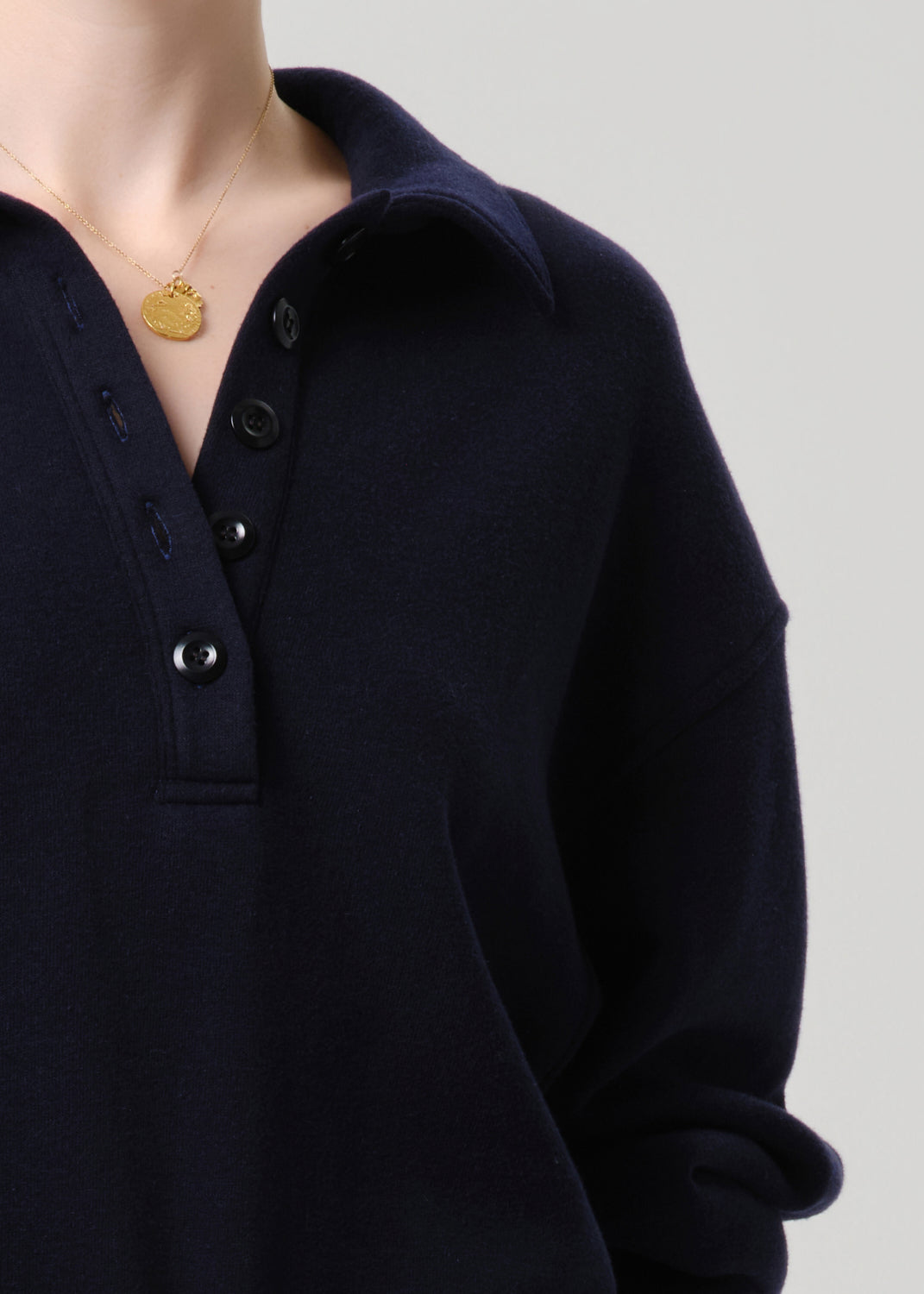 Close-up of a person wearing a dark navy long-sleeve knit polo shirt with a buttoned collar and a gold pendant necklace against a grey background.