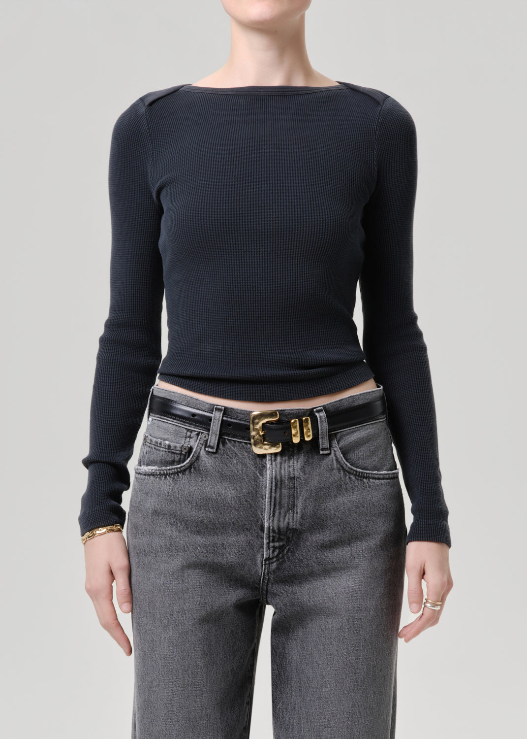 Person wearing a black long-sleeve top and gray jeans with a gold belt on a grey background
