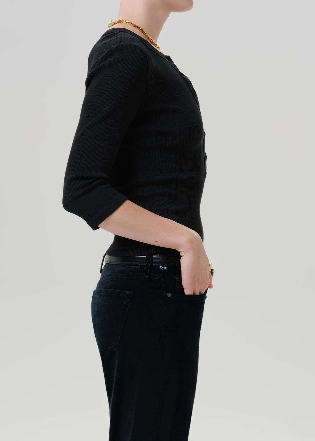 Person wearing a black long sleeve henley top with black pants viewed from the side on a grey background