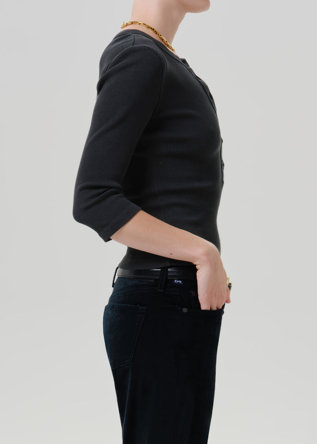 Person wearing a black long sleeve fitted henley top with black jeans, side view on a grey background.