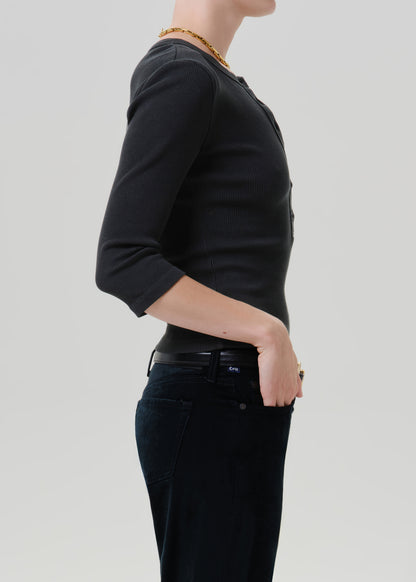 Person wearing a black long sleeve fitted henley top with black jeans, side view on a grey background.